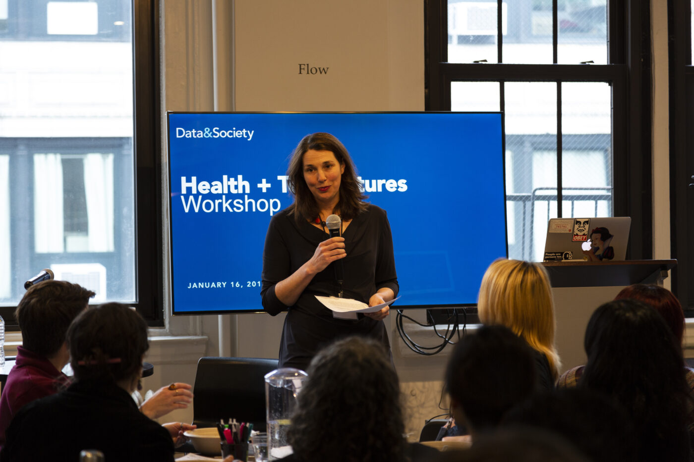 Woman with microphone standing in front of screen that reads Health and Tech Workshop