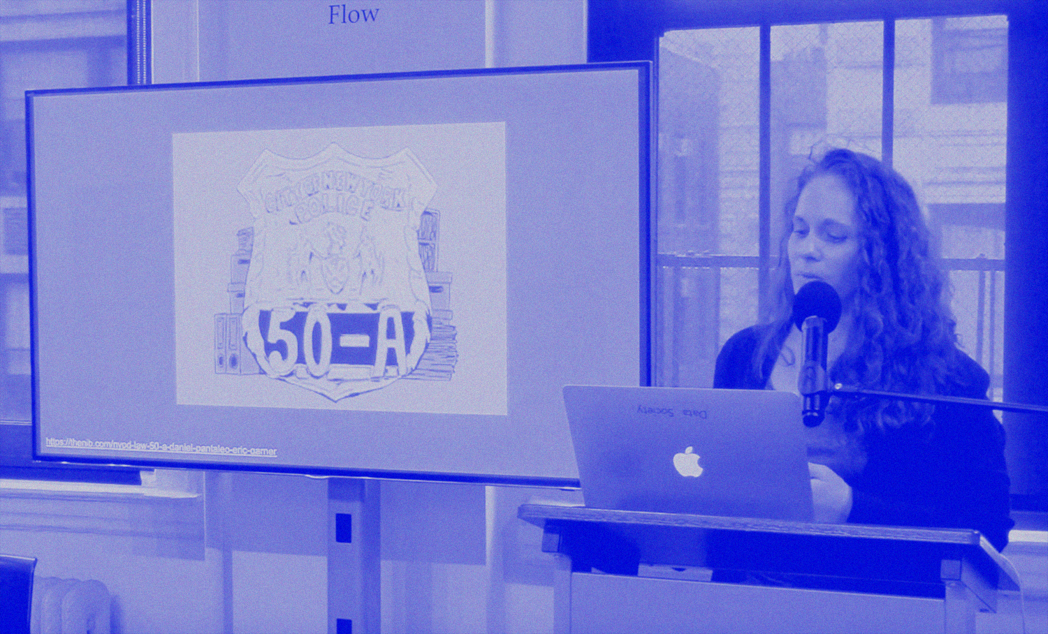 photo of person with long hair speaking into a microphone next to a screen with a large illustration of an NYPD badge with the word 